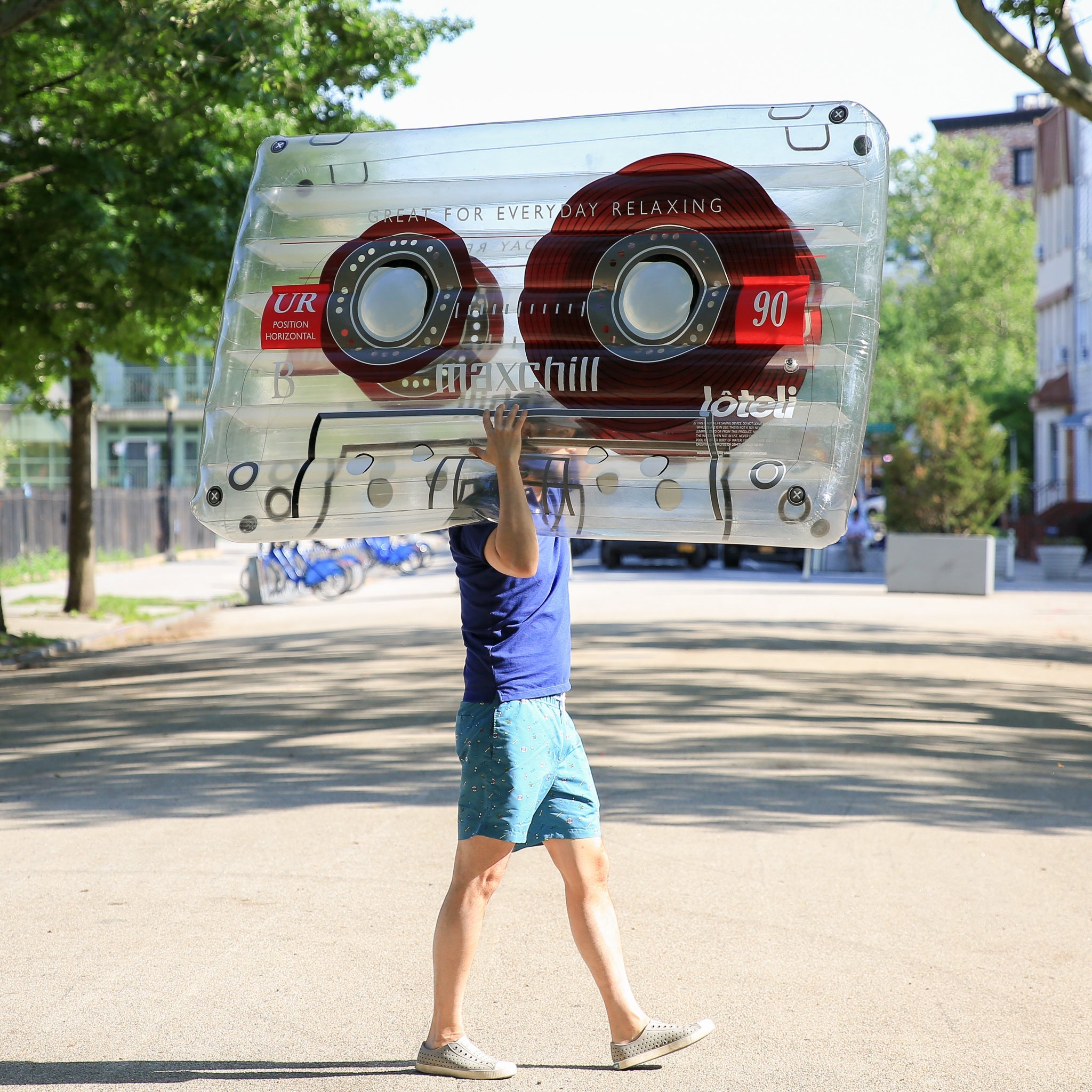 A man carrying a clear cassette float across the street on his near shoulder, which blocks his face from view. The man is wearing blue shorts, blue t shirt and grey sneakers. 