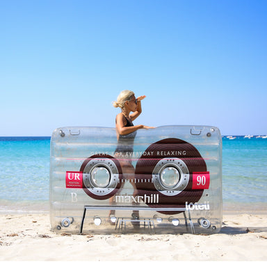 Woman standing behind clear cassette pool float looking to the right with sunglasses on and a hand blocking the sun to look down the coast.  She is standing on the beach and the water is behind her. 