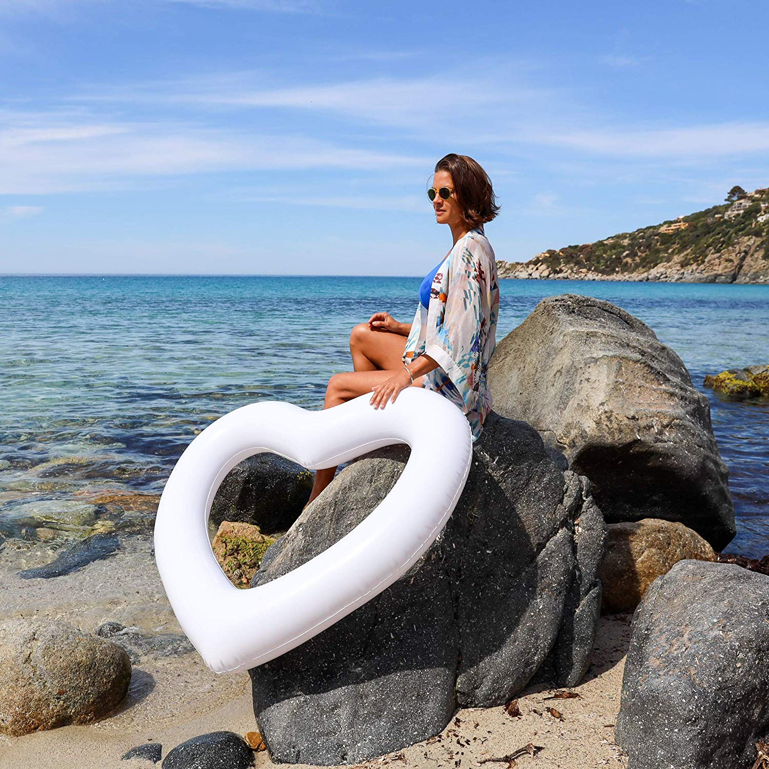 Woman in sunglasses and blue bathing suit sits on the rocks by the beach. She's holding a white heart shaped pool float and looking to the left, head slightly towards the camera but focusing still off in the distance