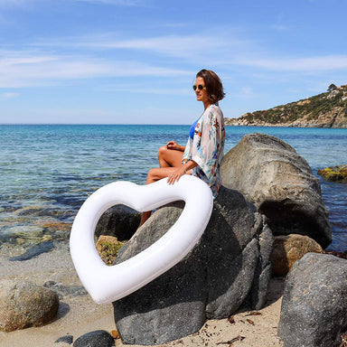Woman in sunglasses and blue bathing suit sits on the rocks by the beach. She's holding a white heart shaped pool float and looking to the left, head slightly towards the camera but focusing still off in the distance