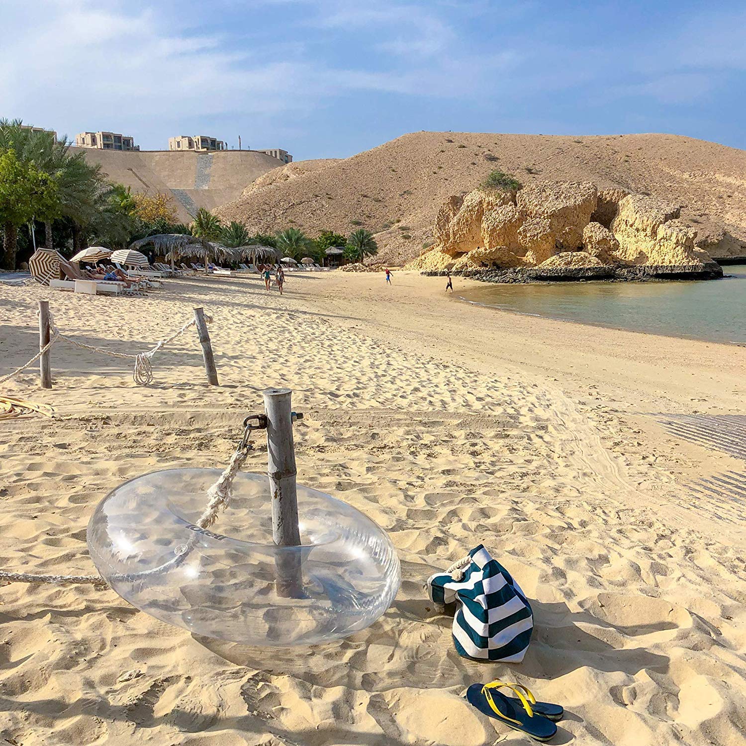 Clear swim ring around a wooden post on the edge of the beach near flip flops and beach bag.
