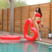 Woman in red swimsuit walks to the right with red heart shaped float in hand, looking twoards pool and away from camera