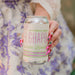 Manicured Hand holding a can of 'El Guapo' Cucumber Lavender Bitters & Soda with a blurred floral background