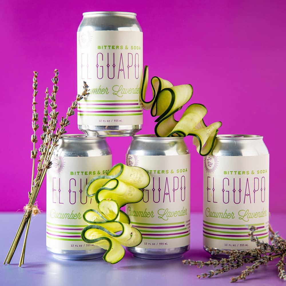 Four cans of El Guapo Cucumber Lavender Bitters & Soda on a purple background with decorative cucumber squigles and dried lavender stalks.