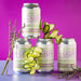 Four cans of El Guapo Cucumber Lavender Bitters & Soda on a purple background with decorative cucumber squigles and dried lavender stalks.