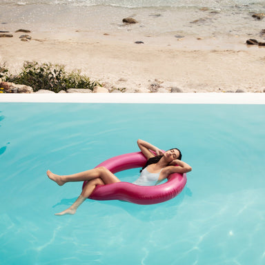 woman in white bathing suit relaxes with her legs crossed and arms behind her head in metallic pink heart shaped float