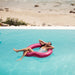 woman in white bathing suit relaxes with her legs crossed and arms behind her head in metallic pink heart shaped float