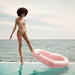 Woman in stylish swimsuit on the edge of infinity pool holds pink heart float up by her foot.