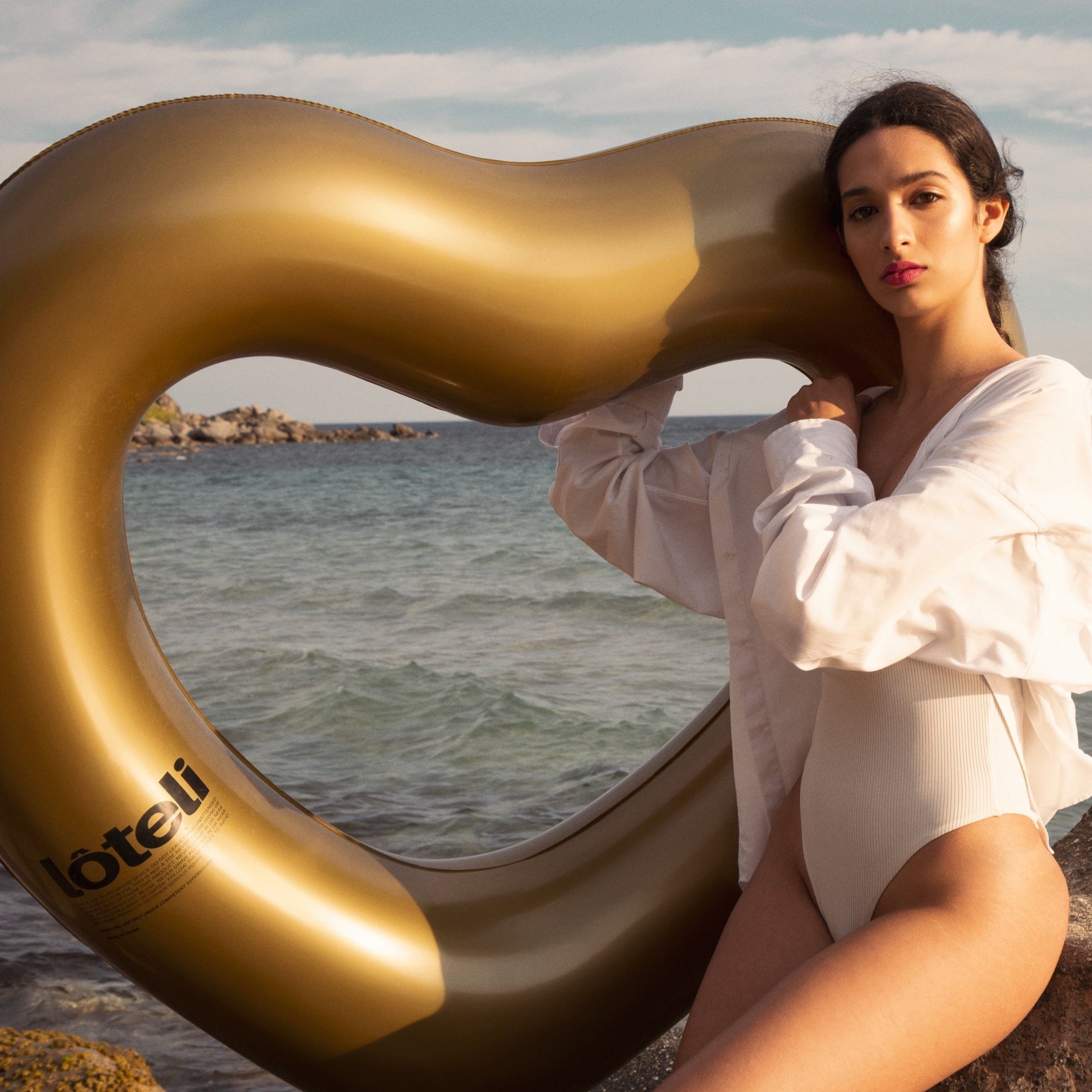 woman resting on rocks by the beach, holding gold heart float behind her. She is facing left and looking at the camera