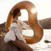 Woman in white is leaning on rocks by the beach, resting gold heart float on the rock with some above her head. She is looking to the right and away from the camera towards the water